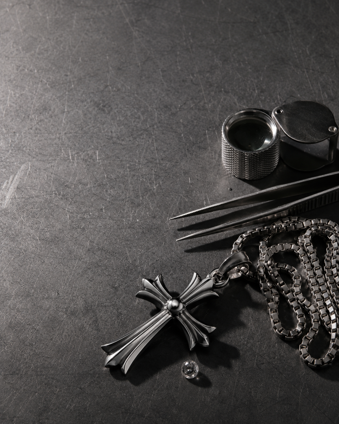 A jeweller's bench with loupe, tweezers, a steel cross pendant, a box chain and a loose stone